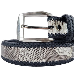 May include: A belt featuring a central panel with a grey and white snakeskin pattern, bordered by dark navy blue braided edges. The belt includes a polished silver-tone metal buckle with a rectangular frame and a single prong for fastening.
