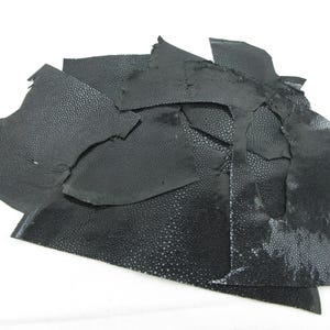 Puede incluir: Una colección de retazos de cuero negro irregulares que presentan una textura granulada y punteada distintiva en toda la superficie. Las piezas varían en tamaño y forma, mostrando un acabado mate con pequeñas protuberancias elevadas.