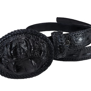 Puede incluir: Un cinturón negro con una hebilla grande de forma ovalada y un patrón texturizado en relieve. La correa del cinturón tiene una textura de escamas rugosas similar a la de un reptil y un detalle de borde trenzado en toda su longitud.