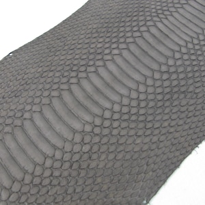 May include: A dark grey material featuring a textured, repeating scale pattern that mimics the appearance of snake skin. The surface displays a central row of elongated, rectangular scales flanked by smaller, rounded scales on both sides.