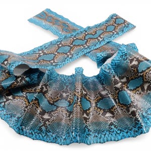 May include: A piece of textured snakeskin leather featuring a vibrant blue, black, and brown diamond-shaped scale pattern. The material is folded and draped, showcasing the detailed organic texture of the scales across the entire surface area.