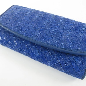 May include: A rectangular clutch wallet in a vibrant royal blue colour. The exterior features a woven pattern constructed from textured, granular material with a smooth blue leather trim along the edges of the flap and the main body of the wallet.