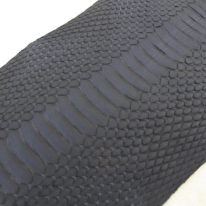 May include: A dark charcoal grey material featuring a textured snakeskin pattern. The surface displays a repeating arrangement of small, rounded scales transitioning into a central row of larger, elongated, rectangular scales running diagonally.