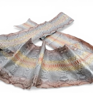 Pode incluir: Uma pele de cobra curtida e plana com um padrão de escamas natural em tons de cinza, marrom e amarelo pálido. A pele é alongada e flexível, exibindo a textura distinta e as escamas sobrepostas características do couro de réptil.