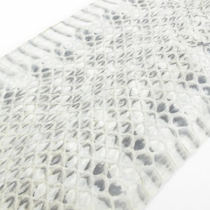 Puede incluir: Una pieza de material plana y rectangular que presenta un patrón de escamas texturizado y repetitivo en tonos blanco, gris claro y carbón. La superficie muestra una cuadrícula orgánica en forma de diamante que imita la piel de reptil.