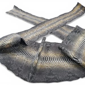 Puede incluir: Una tira larga y flexible de piel de serpiente con un patrón de escamas natural en tonos gris carbón, negro y dorado apagado. El material está dispuesto en una formación cruzada y superpuesta, mostrando la textura detallada de las escamas.