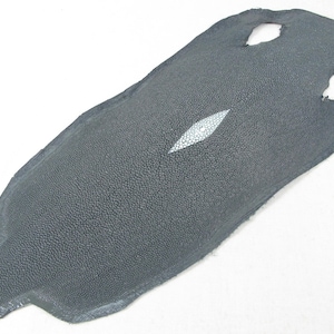 May include: A grey stingray skin featuring a textured, pebbled surface and a distinct, elongated white diamond-shaped calcified centre. The hide has an irregular, tapered shape with two circular cutouts near the top edge of the material.