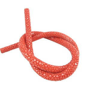 May include: A flexible, cylindrical cord featuring a vibrant red stingray leather texture. The surface is covered in a dense, pebbled pattern of small, rounded, bead-like bumps that create a distinct, tactile, and organic appearance throughout.