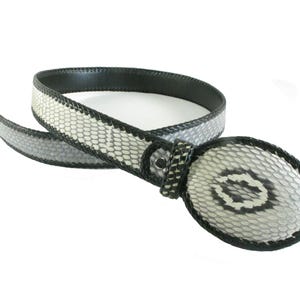 Puede incluir: Un cinturón con un patrón texturizado similar a escamas en tonos blanco, gris y negro. El cinturón incluye una hebilla ovalada a juego con un patrón oscuro central, acabado con bordes cosidos en negro a lo largo de la correa y el perímetro.