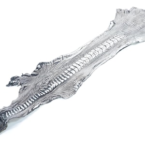 Puede incluir: Una pieza de material plateado, alargada y texturizada, que se asemeja a una piel de reptil. Presenta un patrón central prominente y elevado de columna vertebral con escamas rectangulares segmentadas, terminando en una punta en forma de garra.