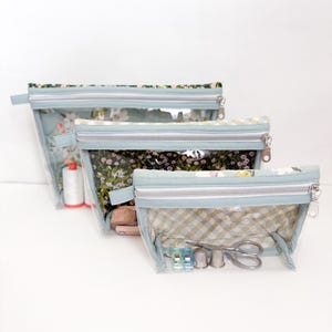 Pode incluir: Três bolsas transparentes com zíper de tamanhos variados. As bolsas têm acabamento em azul claro e detalhes em tecido floral ou xadrez. A menor bolsa contém materiais de costura, incluindo tesouras e dedais. As bolsas estão dispostas em fila.