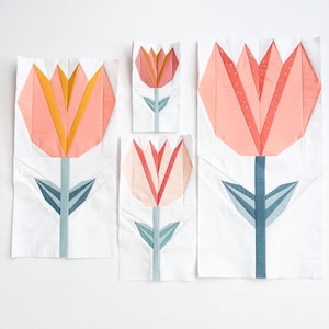 Puede incluir: Cinco bloques de tela con diseños de tulipanes. Los tulipanes tienen pétalos melocotón y amarillos, tallos azul grisáceo y hojas azul grisáceo. El fondo es blanco. Los bloques varían en tamaño, el más grande mide unos 30 cm de alto.