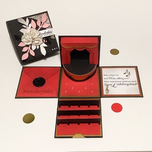 Könnte beinhalten: Eine schwarze und rote Explosionsbox mit einem Bühnen-Design. Die Box ist mit Papierblumen und dem Text "Wunderbar" auf dem Deckel verziert. Im Inneren der Box befinden sich eine Bühne, ein roter Umschlag mit dem Text "Wunscherfüller" und eine Tafel mit Noten und Text.