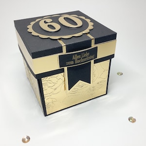 Könnte beinhalten: Schwarze und goldene dekorative Box mit der Zahl 60 auf dem Deckel. Die Box hat goldene Akzente und den Text "Alles Liebe zum Hochzeitstag". Die Box ist quadratisch und hat ein florales Design.