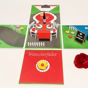 Könnte beinhalten: Eine handgefertigte, mehrteilige Karte mit einer Miniatur-Essszene im Freien. Die Karte zeigt eine rote Tischdecke, weiße Stühle und eine Weinflasche. Die Karte enthält auch ein kleines Tafelschild mit der Aufschrift "Heute geöffnet". Eine rote Papierrose ist ebenfalls enthalten.