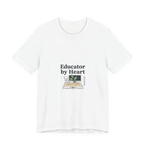 May include: White t-shirt with the text "Educator by Heart" in black. The design includes a book, computer, pencil, and plant illustration. The shirt is made of a soft material.