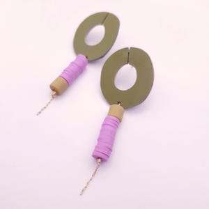 Può includere: Un paio di orecchini fatti a mano con forme ovali verde oliva con una fessura nella parte superiore. Gli orecchini hanno un cilindro lavanda con un accento beige e una catena color oro. Su sfondo bianco.