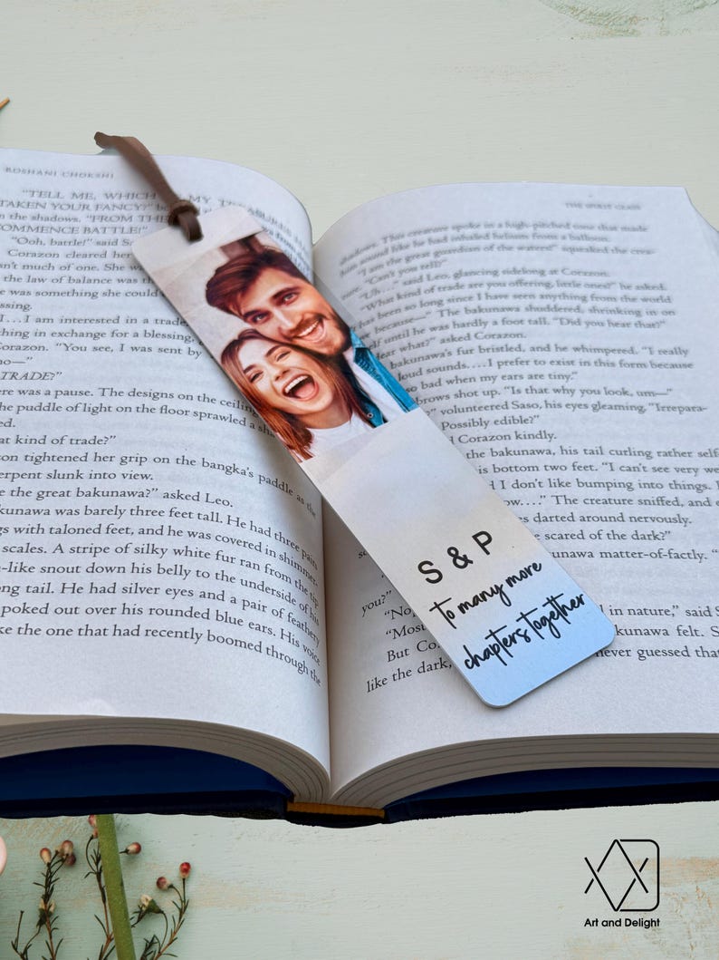 May include: A silver bookmark with a photo of a couple smiling. The bookmark has the text "S&P To many more chapters together" printed on it.