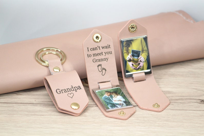 K&ouml;nnte beinhalten: Hellrosa Leder-Schl&uuml;sselanh&auml;nger mit goldenen Beschl&auml;gen. Ein Schl&uuml;sselanh&auml;nger mit der Aufschrift "Grandpa" mit Herz, ein weiterer mit "I can't wait to meet you Granny" mit Babyf&uuml;&szlig;en und der dritte mit einem Ultraschallbild.