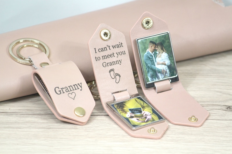 May include: Pink leather keychains with metal key rings. One keychain has the word "Granny" and a heart. Another keychain has the text "I can't wait to meet you Granny" with baby footprints. Both keychains have a photo inside.