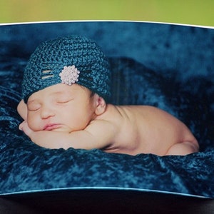Pode incluir: Um bebê dormindo usando um chapéu azul de crochê com um enfeite de flor branca, deitado em uma superfície de veludo azul.