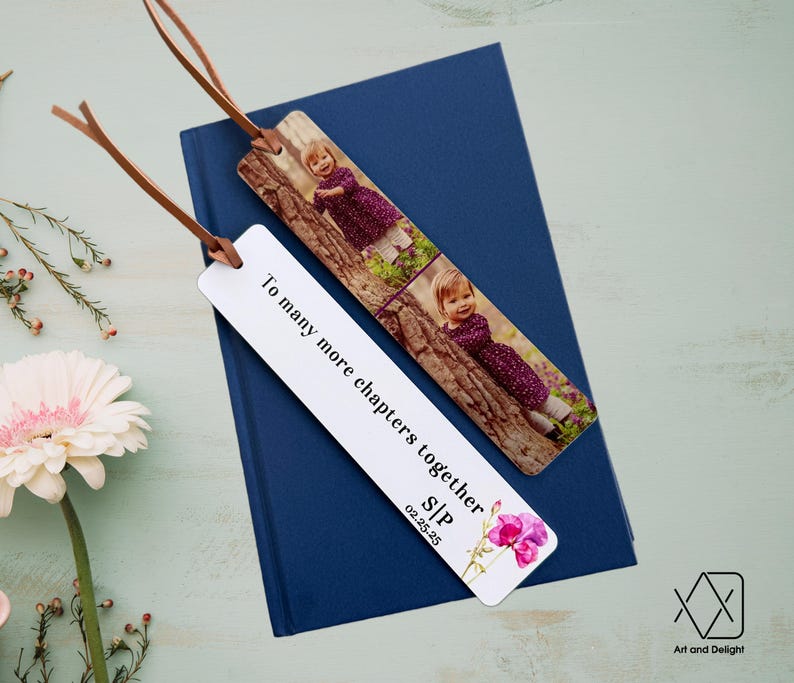 May include: White bookmark with the text "To many more chapters together SIP 02.25.25" and a watercolor illustration of pink flowers. The bookmark has a photo of a young girl in a purple dress on a tree branch.