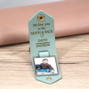 K&ouml;nnte beinhalten: Hellblaues Leder-Schl&uuml;sselanh&auml;nger mit einem Foto eines Kindes. Der Schl&uuml;sselanh&auml;nger tr&auml;gt den Text "We love you to the MOON & BACK" und die Namen "DAVID" und "ANDREW". Er wird mit einem goldenen Druckknopf verschlossen.