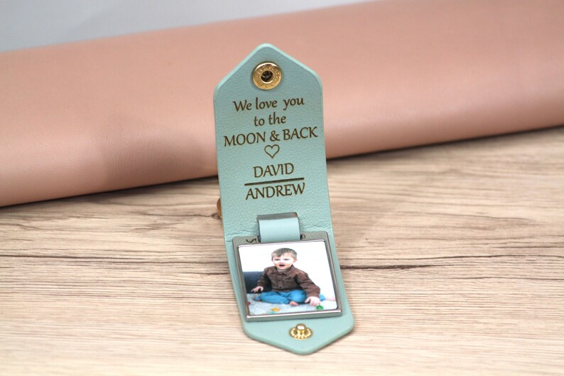 K&ouml;nnte beinhalten: Hellblaues Leder-Schl&uuml;sselanh&auml;nger mit einem Foto eines Kindes. Der Schl&uuml;sselanh&auml;nger tr&auml;gt den Text "We love you to the MOON & BACK" und die Namen "DAVID" und "ANDREW". Er wird mit einem goldenen Druckknopf verschlossen.