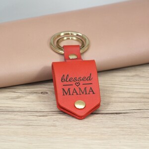 May include: A red leather keychain with a gold key ring. The keychain has the words "blessed MAMA" engraved on it. The keychain is on a light wood surface.