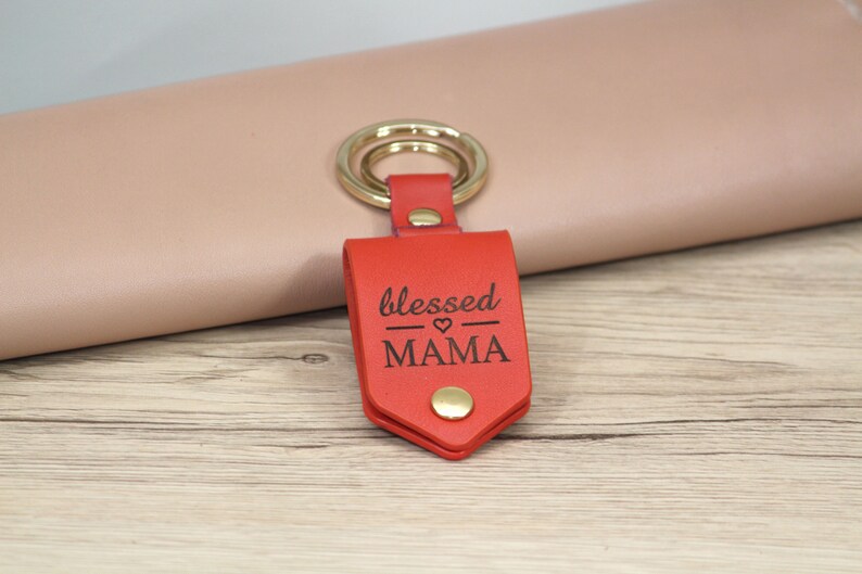 May include: A red leather keychain with a gold key ring. The keychain has the words "blessed MAMA" engraved on it. The keychain is on a light wood surface.