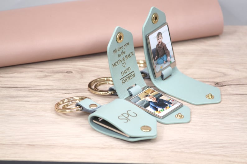 K&ouml;nnte beinhalten: Hellblaue Leder-Schl&uuml;sselanh&auml;nger mit goldfarbener Hardware. Jeder Schl&uuml;sselanh&auml;nger zeigt ein Foto und personalisierten Text: "We love you to the MOON & BACK" und Namen. Ein Schl&uuml;sselanh&auml;nger hat die Initialen "SPG". Die Schl&uuml;sselanh&auml;nger liegen auf einer Holzoberfl&auml;che.