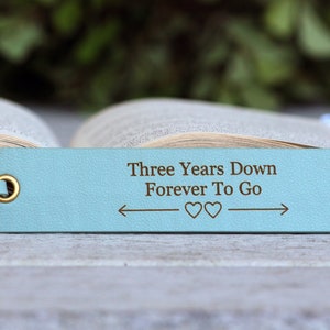 May include: A light blue leather bookmark with the text "Three Years Down Forever To Go" and two hearts. The date "9-15-2020" is also engraved on the bookmark.