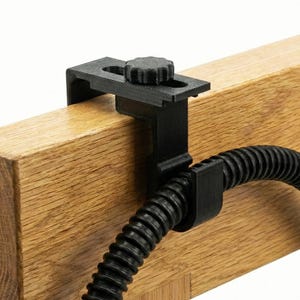 Op de afbeelding: Zwarte 3D-geprinte klem met een knop, bevestigd aan een houten balk. De klem houdt een zwarte, flexibele, geribbelde buis vast. De houtnerf is zichtbaar op de balk.