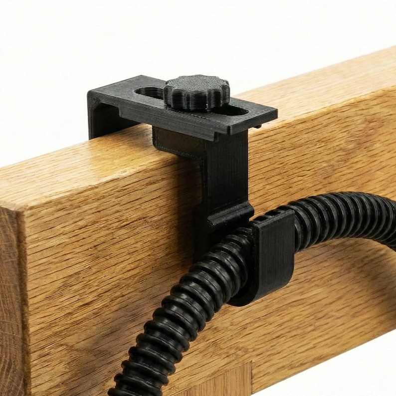 Op de afbeelding: Zwarte 3D-geprinte klem met een knop, bevestigd aan een houten balk. De klem houdt een zwarte, flexibele, geribbelde buis vast. De houtnerf is zichtbaar op de balk.