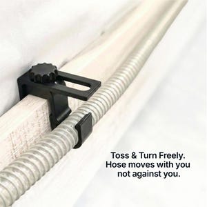 Op de afbeelding: Zwarte slanghouder bevestigd aan een houten oppervlak, met een flexibele zilveren slang die erdoorheen loopt. De houder heeft een knop voor afstelling en de tekst "Toss & Turn Freely. Hose moves with you not against you."