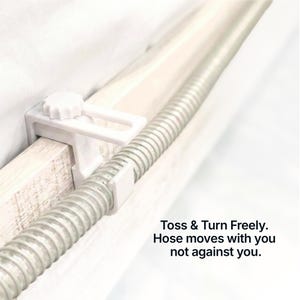 Op de afbeelding: Een witte plastic slanghouder met een knop, bevestigd aan een houten frame. Een flexibele, zilveren slang wordt op zijn plaats gehouden door de houder. Tekst op de afbeelding luidt: Toss & Turn Freely. Hose moves with you not against you.