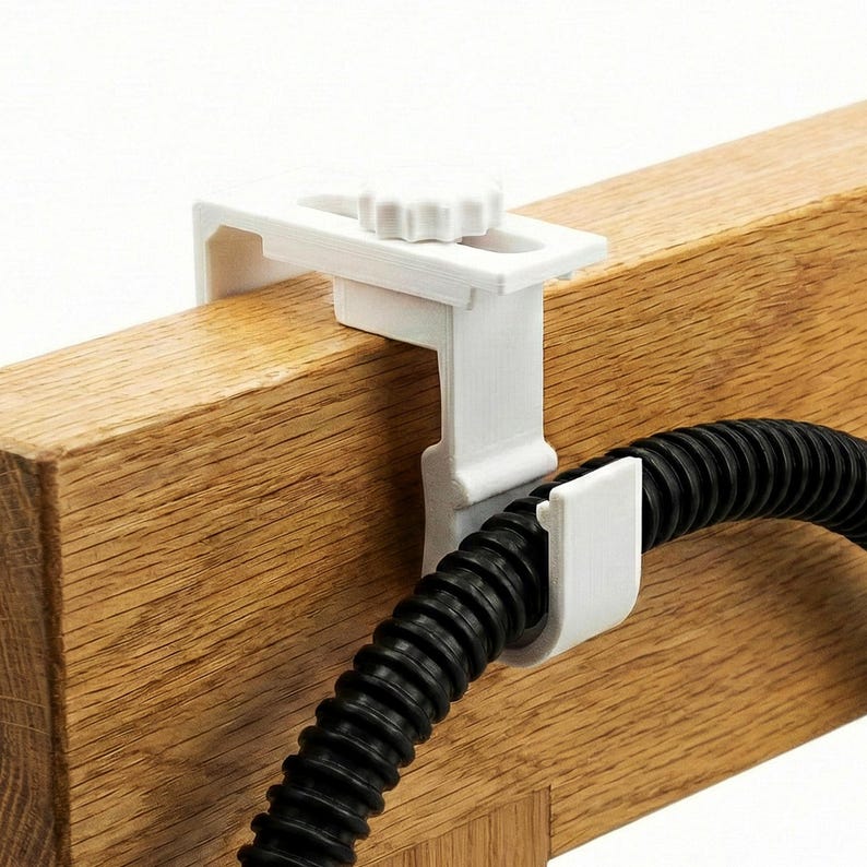 Op de afbeelding: Een witte plastic klem met een knop, bevestigd aan een houten balk, die een zwarte gegolfde slang vasthoudt. De klem heeft een rechthoekig ontwerp met een gebogen gedeelte om de slang vast te zetten. Het hout heeft een natuurlijke nerf.