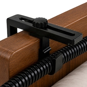 May include: A black plastic adjustable clamp mounted onto a brown wood-grain surface. The clamp features a horizontal slotted arm with a circular adjustment knob and is connected to a black flexible corrugated tube running horizontally.