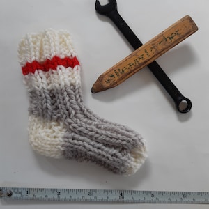 May include: A gray and white knitted sock with a red stripe, a wooden pencil with a green label, and a black wrench on a white background.