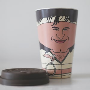 May include: A brown and black reusable coffee cup with a cartoon illustration of a person with brown hair wearing a white headband and a white shirt with a green collar. The person is holding a tennis racket. The cup has a matching brown lid.
