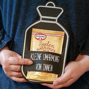 May include: A black and white cardboard cutout of a bottle with a yellow packet of Dr. Oetker "Seelenwärmer" soup mix inside. The bottle has the text "Kleine Umarmung von Innen" on it.