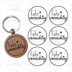 Könnte beinhalten: Ein Holzschlüsselanhänger mit einem schwarzen Kreis und dem Text "Fahr vorsichtig" mit einem kleinen Herzsymbol. Der Schlüsselanhänger ist von fünf weiteren schwarzen Kreisen mit dem gleichen Text und Herzsymbol umgeben.