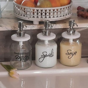 Könnte beinhalten: Drei Seifenspender aus Glas mit silbernen Pumpen und weißen Deckeln. Die erste Flasche ist leer und mit Desinfektions Mittel beschriftet, die zweite enthält weiße Flüssigkeit mit der Aufschrift Spüli und die dritte gelbe Flüssigkeit mit der Aufschrift Seife.