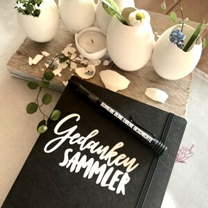 Könnte beinhalten: Ein schwarzes, ledergebundenes Tagebuch mit den Worten "Gedanken Sammler" in weißer Schrift. Ein schwarzer Stift mit den Worten "Schreibe deine eigene Geschichte" liegt auf dem Tagebuch. Das Tagebuch ist von weißen Eierschalen mit Blumen darin umgeben.