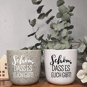 Könnte beinhalten: Zwei Keramikbecher mit gesprenkeltem Überzug. Ein Becher ist grau mit weißem Text, der "Schön dass es euch gibt!" lautet. Der andere Becher ist weiß mit schwarzem Text, der "Schön dass es euch gibt!" lautet.