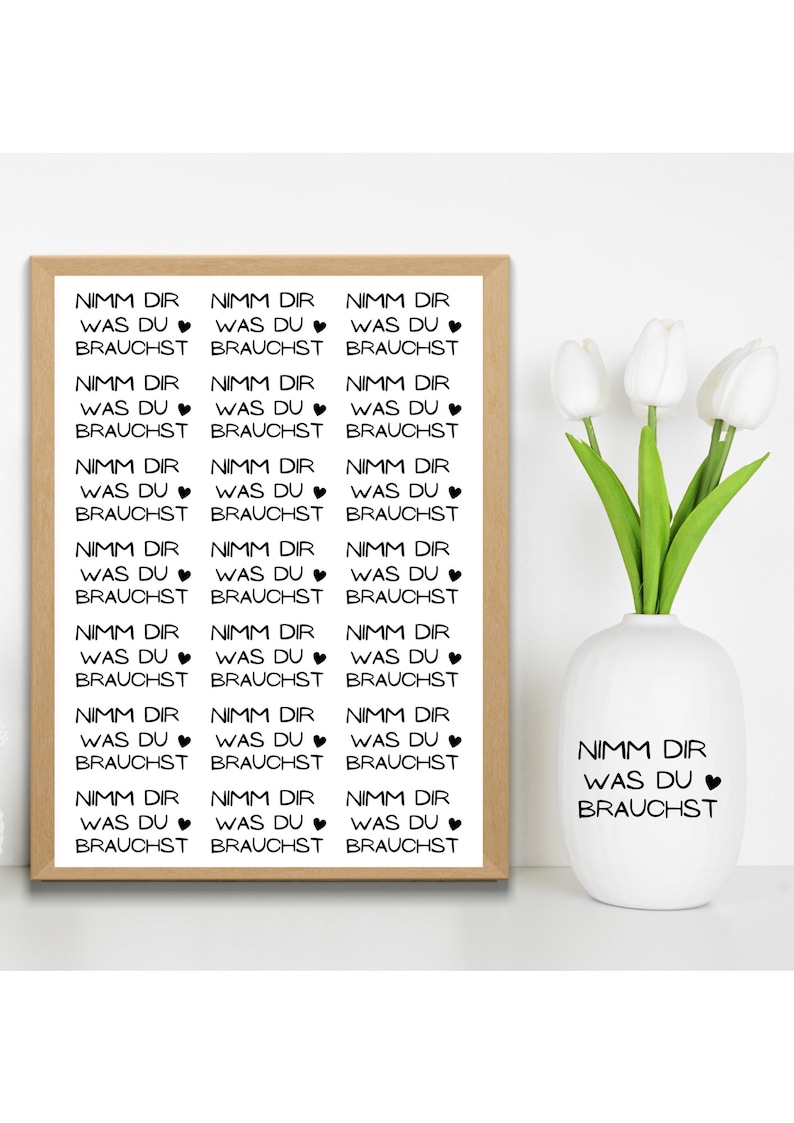 May include: A framed print with the German text "Nimm dir was du brauchst" in a repeating pattern. A white vase with the same text and a heart symbol is next to the print. White tulips are in the vase.