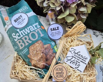 May include: A gift basket with a bag of Schwarz Brot, a small glass vial of salt, a wooden keychain with "Home Sweet Home" text, and a card with German text. The basket is filled with straw and decorated with flowers.
