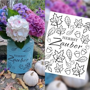 Könnte beinhalten: Eine hellblaue Metallvase mit einem Blumenarrangement aus lila, rosa und weißen Blumen. Die Vase trägt die Worte "HERBST Zauber" und Blatt- und Eicheldesign. Zwei weiße Kürbisse und Herbstlaub im Vordergrund.