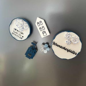 May include: A collection of decorative magnets in various shapes and designs. Includes round magnets with floral accents and text, a house-shaped magnet, and a flower-shaped magnet. The color palette features white, blue, and a touch of silver.