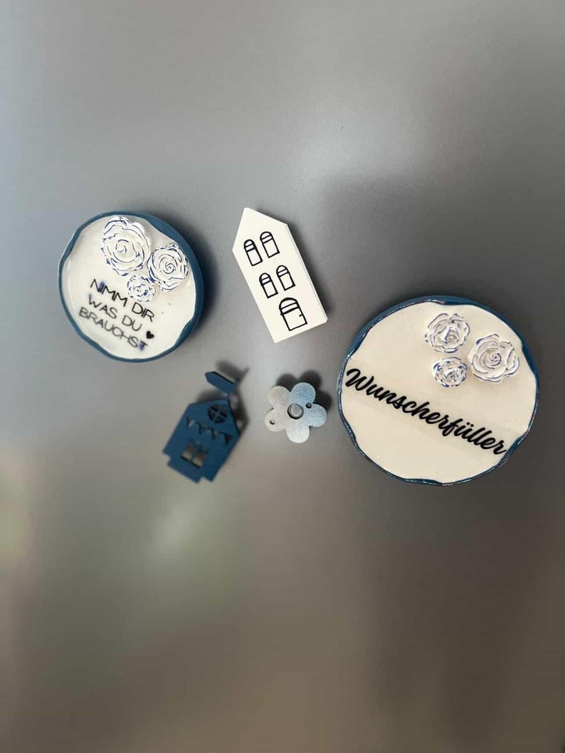 May include: A collection of decorative magnets in various shapes and designs. Includes round magnets with floral accents and text, a house-shaped magnet, and a flower-shaped magnet. The color palette features white, blue, and a touch of silver.